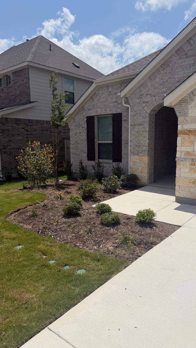 Modern two-story brick and stone house with dark shutters, manicured front landscaping, concrete walkway, and blue cloudy sky